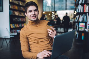 Modern kütüphanede dizüstü bilgisayarı incelerken gülen ve bir fincan sıcak kahve içen neşeli bir adam.