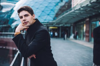 Siyah ceketli, çenesine dokunan ve gündüz vakti sokakta ayakta duran ciddi bir erkeğin yan görüntüsü.