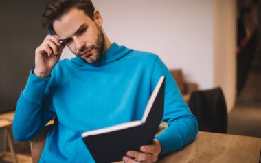 Mavi kapüşonlu beyaz bir genç üniversitede ilginç edebiyat kitapları okuyor. Sıradan giyinmiş zeki bir erkek öğrenci kapalı alanda eğitim kitaplarından öğrenim görüyor.