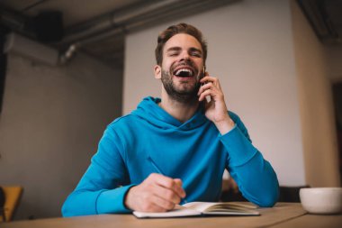 Neşeli erkek öğrenci, eğitim kitabındaki sınav notları hakkında konuşmak için üniversiteden profesöre telefon ediyor. Heyecanlı hipster adam cep telefonuyla konuşuyor ve içeride gülüyor.