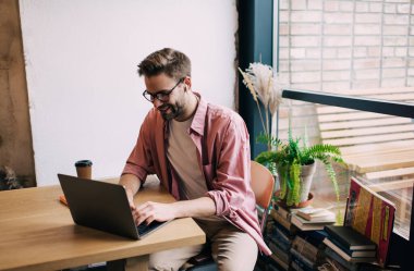 Klasik gözlüklü ve bluetooth kulaklıklı mutlu beyaz erkek modern dizüstü bilgisayarda web sitesine göz atıyor, neşeli bilişim uzmanı dijital netbook 'ta medya uygulaması aracılığıyla grafik tasarım yaratıyor.
