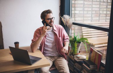 Uzaktan kumandalı iş sırasında cep telefonuyla sohbet etmekten hoşlanan mutlu erkek serbest yazar. Gözlüklü neşeli hippi dizüstü bilgisayarla masaya gülümsüyor.