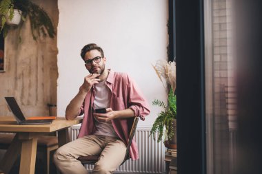 Görme düzeltmesi için optik gözlüklü düşünceli bir erkek akıllı telefon aleti tutuyor ve düşünüyor. Elektronik stilus mobil cihazı ve dizüstü bilgisayarı olan düşünceli bir hipster.