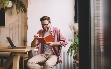 Uzaktan kumandalı kahve molası sırasında gülümseyen edebiyatı olan mutlu adam bulanık netbook üzerinde çalışıyor, en çok satanlar kitabını tutan neşeli erkek öğrencinin portresi.