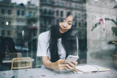 Gündüz vakti modern beyaz cep telefonuyla sörf yapan, uzun siyah saçlı, çağdaş şehir kafeteryasından genç Asyalı bayana bakın.