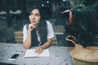 Düşünceli, Asyalı, uzun saçlı, kadın yazar, kafeteryada deftere yazı yazarken elini uzatıp gözlerini kaçırıyor.