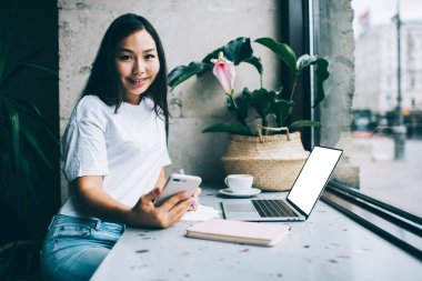 Kafede dizüstü bilgisayarını açıp mesajlaşırken ve not alırken kameraya bakan mutlu Asyalı genç kız öğrenci.