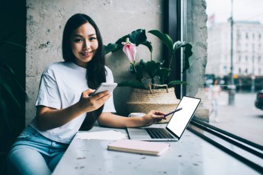 Neşeli, Asyalı, sıradan giyinen genç bir kız öğrenci akıllı telefondan mesajlaşırken ve kameraya bakarken dizüstü bilgisayarına kalemle işaret ediyor.