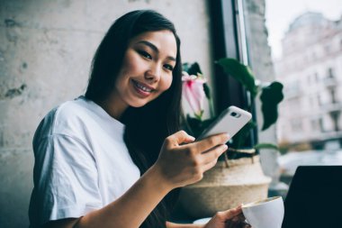 Mutlu genç Asyalı kadın seramik beyaz bardakta sıcak aromatik içecek içiyor ve akıllı telefondan mesajlaşırken gülümseyen kameraya bakıyor.