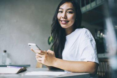 Genç Asyalı kız öğrenci kameraya bakarken akıllı telefondan mesaj atıyor ve kafede oturan ajandaya not alıyor.