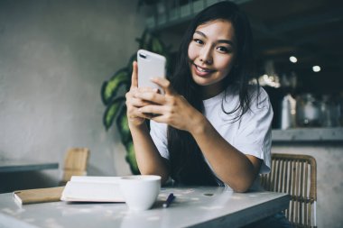 Positif Asyalı bir kadın cep telefonuyla mesajlaşırken ve arka plandaki bulanık kameraya bakarken net kitaplar ve bir bardak içkiyle masada oturuyor.