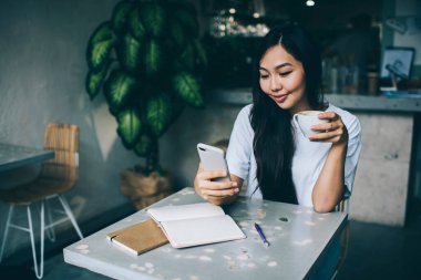 Kafeteryadaki not defterleriyle masada otururken bir fincan sıcak kahve içen neşeli Asyalı kadın.