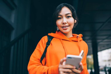 Sokakta cep telefonuna bakarken, koyu renk saçlı, parlak günlük kıyafetli, pozitif Asyalı kız öğrenci kameraya bakıyor.