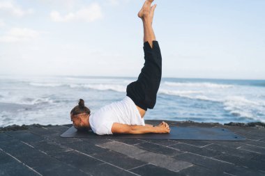 Spor giyim antrenmanındaki genç erkek atlet, sabah antrenmanında asana solunumunu dengeliyor ve ilham alıyor, beyaz adam yoga yaparken deniz kenarındaki sette vücut kaslarını esnetiyor.