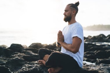 Nilüfer çiçeğinde oturan ve namaste pozu veren sportif giyimli bir erkek okyanus kıyısında sabah meditasyonu sırasında dua ederken, yoga konsantrasyonu sırasında gözleri kapalı olan genç beyaz bir adam doğada dinleniyor.
