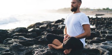 Kıyı şeridinde dinlenen genç adam meditasyon ve iyileşme için gündüzleri bütünsel inzivaya çekiliyor. Pranayama asana 'da spor giysileri içinde nefes alan beyaz erkek deniz kıyısında hatha antrenmanı yapıyor.