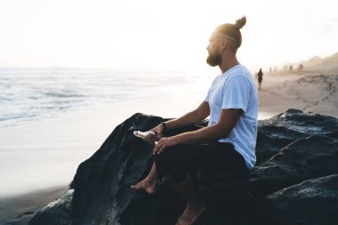 Sahil şeridindeki Rosky Stone 'da dinlenen erkek turistin yan görüntüsü ve boş zamanlarında vahşi sahili gezmek için deniz manzarası, hafta sonu tatillerinde serbest kıyafet giyen genç adam.
