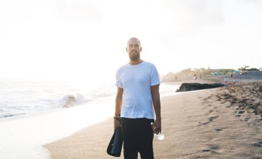 Haftasonu deniz kıyısında esnemek ve poz vermek için elinde bir şişe su ve minder tutan yarı uzunluğunda sportif bir adam. Kafkas erkek yogi 'nin boş zamanlarını kumlu sahil şeridinde geçirdiği portresi.