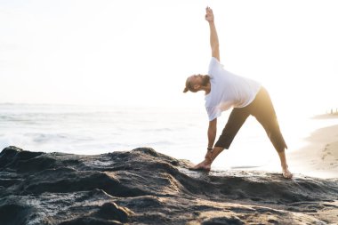 Beyaz erkek yogi kıyı şeridi boyunca esneme ve antrenman yapıyor. Haftasonunu bütünsel iyileşme için geçiriyor. Genç adam zindelik ve sağlıklı yaşam tarzına ulaşmak için egzersiz yapıyor.