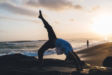 Esnek erkek sporcu sabah pilates antrenmanı sırasında kaslarını esneterek okyanus kenarında spor kıyafetleri giymiş, yoga asanası yapan ve sağlıklı yaşam tarzını korumak ve bütünsel canlılığa ulaşmak için yoga yapan beyaz bir adam.
