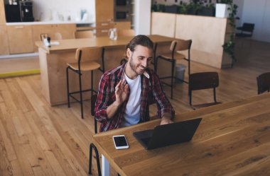 Milenyum Kafkas işadamı laptopta video görüşmesi sırasında el sallıyor. Modern başarılı adam kavramı. Genç gülümseyen erkek girişimci açık uzay ofisindeki masada oturuyor ve çalışıyor.