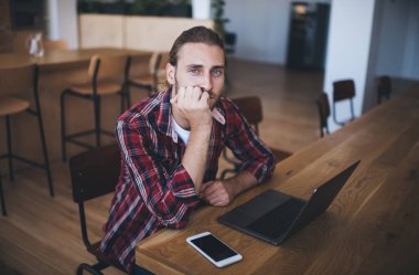 Milenyum Kafkas işadamı dizüstü bilgisayarda çalışırken kameraya bakıyor. Modern başarılı adam kavramı. Genç gülümseyen erkek girişimci açık uzay ofisindeki ahşap masada oturuyor.