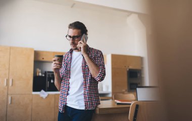 Milenyum Kafkasyalı iş adamı kahve içerken iş sırasında akıllı telefondan konuşuyor. Modern başarılı adam kavramı. Gözlüklü genç ciddi girişimci açık uzay ofisindeki ahşap masada duruyor.