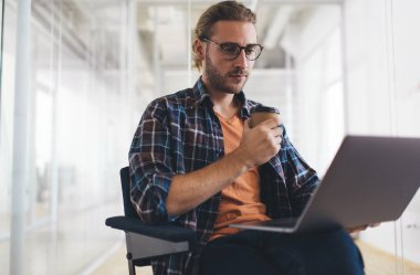 Milenyum Kafkas işadamı elinde kahveyle dizüstü bilgisayardan izliyor. Modern başarılı adam kavramı. Gözlüklü genç girişimci ofiste oturuyor ve ofis koltuğunda çalışıyor.