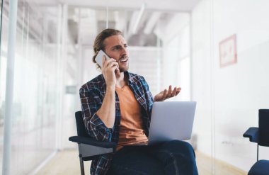 Bin yıllık Kafkasyalı iş adamı dizüstü bilgisayarla çalışırken akıllı telefondan konuşuyor. Modern başarılı adam kavramı. Açık uzay ofisindeki ofis koltuğunda oturan genç girişimci.