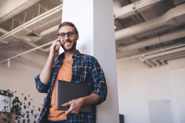 Bin yıllık beyaz iş adamının bilgisayarlı akıllı telefonuyla konuşmasının alt görüntüsü. Modern başarılı adam kavramı. Genç sakallı, gülümseyen, gözlüklü, ofiste kameraya bakan bir girişimci.