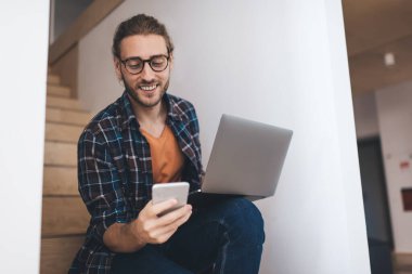 Milenyum Kafkasyalı iş adamı iş sırasında akıllı telefon kullanıyor. Modern başarılı adam kavramı. Genç, yakışıklı, güler yüzlü, gözlüklü, iş yerinde merdivenlerde oturan bir girişimci.