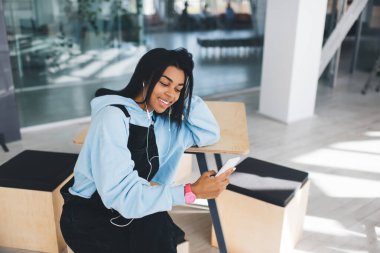 Zenci kız kulaklıkla müzik dinliyor ve cep telefonundan bir şeyler izliyor. Modern kadın yaşam tarzı kavramı. Genç, gülümseyen bir kadın tahta masada oturuyor. Güneşli ofisin içi bulanık