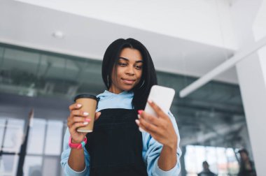 Akıllı telefon kullanan ve kahve içen Afrikalı bir kız. Modern kadın yaşam tarzı kavramı. Sıradan kıyafetler ve kol saati takan gülümseyen genç bir bayan. Modern ofisin içi bulanık. Alt görünüm