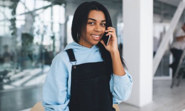 Siyah kız ahşap masada dururken cep telefonuyla konuşuyor. Modern kadın yaşam tarzı kavramı. Gündelik kıyafetler giyen ve kameraya bakan genç bir kadın. Ofisin içi bulanık