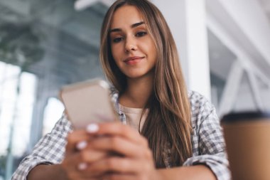 Avrupalı bir kız cep telefonundan bir şey kullanıyor ve izliyor. Modern başarılı kadın kavramı. Sıradan kıyafetler giyen, gülümseyen genç bir kadının seçici odak noktası.