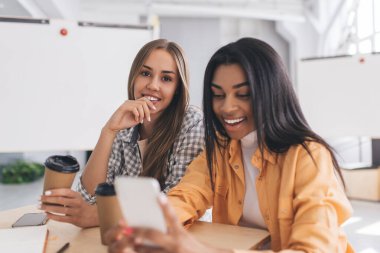 Avrupalı kız arkadaşı kameraya bakarken siyahi kadın cep telefonundan bir şey izliyor. Modern başarılı kadın kavramı. Arkadaşlık ve takım çalışması fikri. Ofisteki masada kahve içen kızlar.