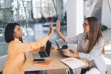 İş kadınları ofiste çalışırken birbirlerine beş dolar veriyorlar. Modern başarılı kadınlar. Takım çalışması kavramı. Masasında ofis malzemeleri, kahve ve aletlerle gülümseyen siyah ve Avrupalı genç kızlar var.