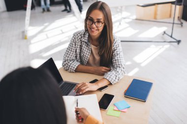Ofiste dizüstü bilgisayar ve defterle çalışan iş kadınları. Modern başarılı kadın kavramı. Takım çalışmasının bir fikri. Ofis malzemeleri ve cep telefonlarıyla masada oturan genç siyahi ve Avrupalı kızlar.