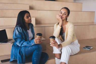 İş kadınları iş sırasında kahve içerken mola veriyorlar. Genç Avrupalı ve siyah kızlar çalışma odasındaki ahşap banklarda. Modern başarılı kadın kavramı. İş başında dinlenme fikri