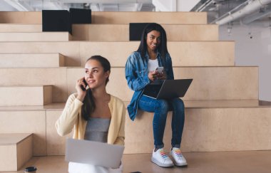 Çalışırken cep telefonu ve laptop kullanan çok ırklı iş kadınları. Modern başarılı kadın kavramı. Genç gülen Avrupalı ve siyah kızlar çalışma odasındaki ahşap banklarda oturuyorlar.