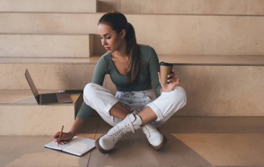 Avrupalı kız deftere bir şeyler yazıyor ve ders sırasında kahve içiyor. Modern başarılı öğrenci kavramı. Genç odaklı kadın çalışma alanında ahşap bankta oturuyor.