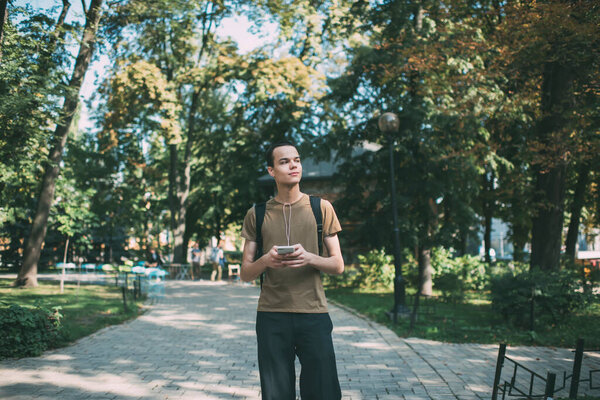 Thoughtful man in casual outfit looking away while standing in park with green trees on sunny day and chatting on mobile phone