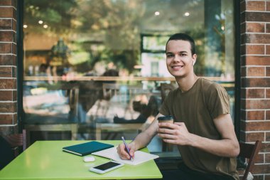 Genç, dişlek bir gülümsemesi ve bir fincan kahvesi olan yakışıklı bir erkek öğrenci kameraya bakıyor ve çalışırken notlar alıyor.