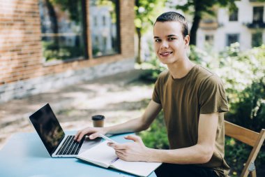 Neşeli, uzaktan kumandalı erkek çalışan, elinde not defteri ve cep telefonuyla sokakta oturmuş iş yaparken kameraya bakıyor.