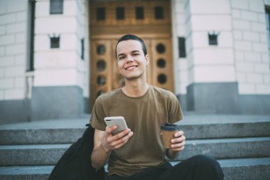 Yüzünde dişlek bir gülümsemeyle merdivenlerde oturmuş elinde bir fincan kahveyle akıllı telefondan sosyal ağları kontrol eden olumlu bir adam.