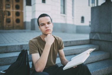Konsantre olmuş erkek öğrenci, elinde çenesiyle merdivenlerde oturmuş ödevlerini yaparken kameraya bakıyor.