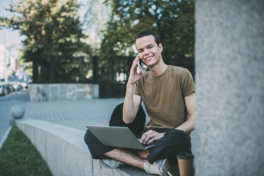 Tüm vücudu telefon görüşmesi yapan ve kameraya bakan mutlu bir erkek. Taş sınırında otururken, arka planda çapraz bacaklı netbook ile.