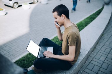 Yukarıdan bakıldığında, odaklanmış bir erkeğin telefon konuşması yapması ve siyah ekranlı bir netbook ile sörf yapması kaldırım kenarındaki taş çitin üzerinde oturması.
