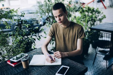 Açık kafeteryadaki tablet ve akıllı telefonla yeşil bitkilerle arka planda bulanık arka planda çalışırken not defterine not alan odaklanmış erkek öğrenci.