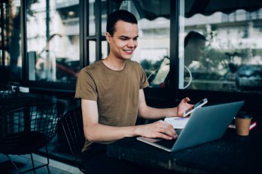 Gündelik giysiler içinde gülümseyen adam masada oturuyor ve kafenin terasında çalışırken akıllı telefon ve laptop kullanıyor.